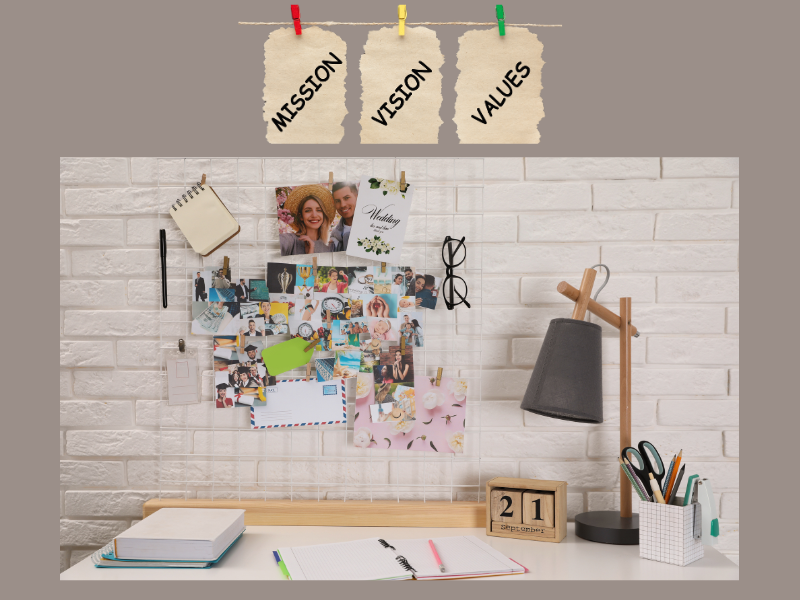 Creative vision board with photos, notes, and sketches pinned to a wall above a desk, symbolizing brainstorming ideas. Three hanging papers labeled "Mission," "Vision," and "Values" emphasize purpose and direction.